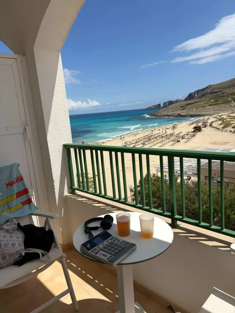 En MPC-sampler och hörlurar på ett vitt bord på en balkong med utsikt över en solig strand och turkost hav. Musikproduktion och semester