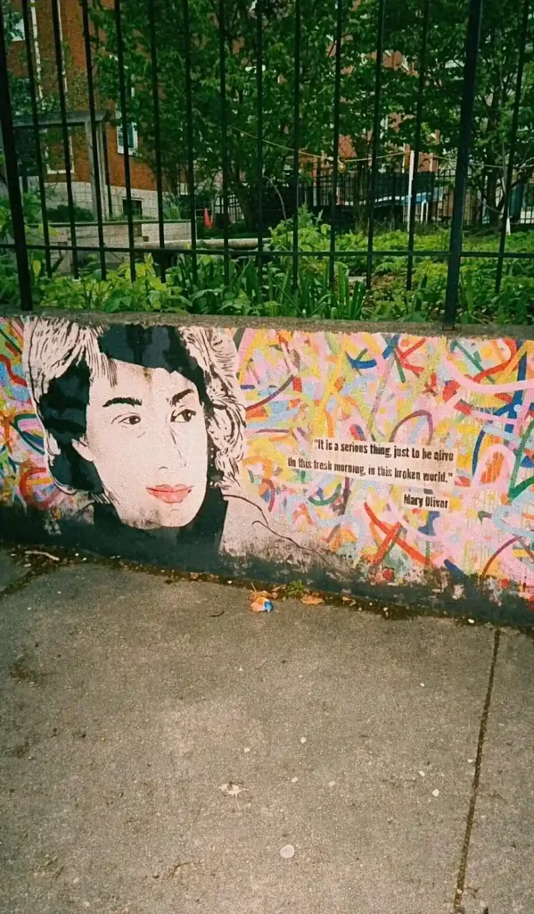 En färgglad väggmålning på en betongmur med ett porträtt av poeten Mary Oliver och ett textcitat bredvid ett svart metallstaket.