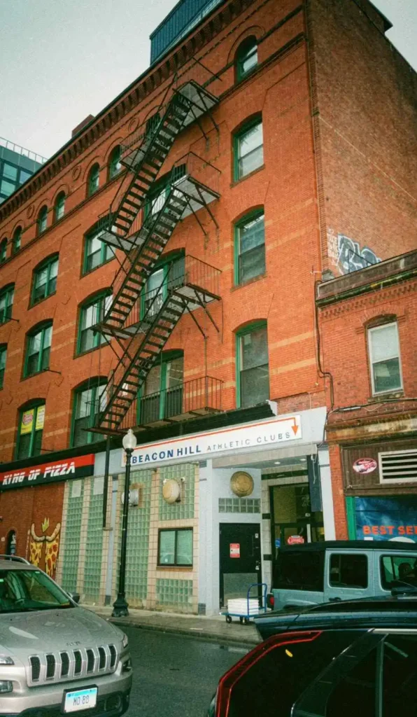En hög röd tegelbyggnad i Boston med en klassisk utvändig brandstege i svart metall på fasaden samt parkerade bilar på gatan nedanför.