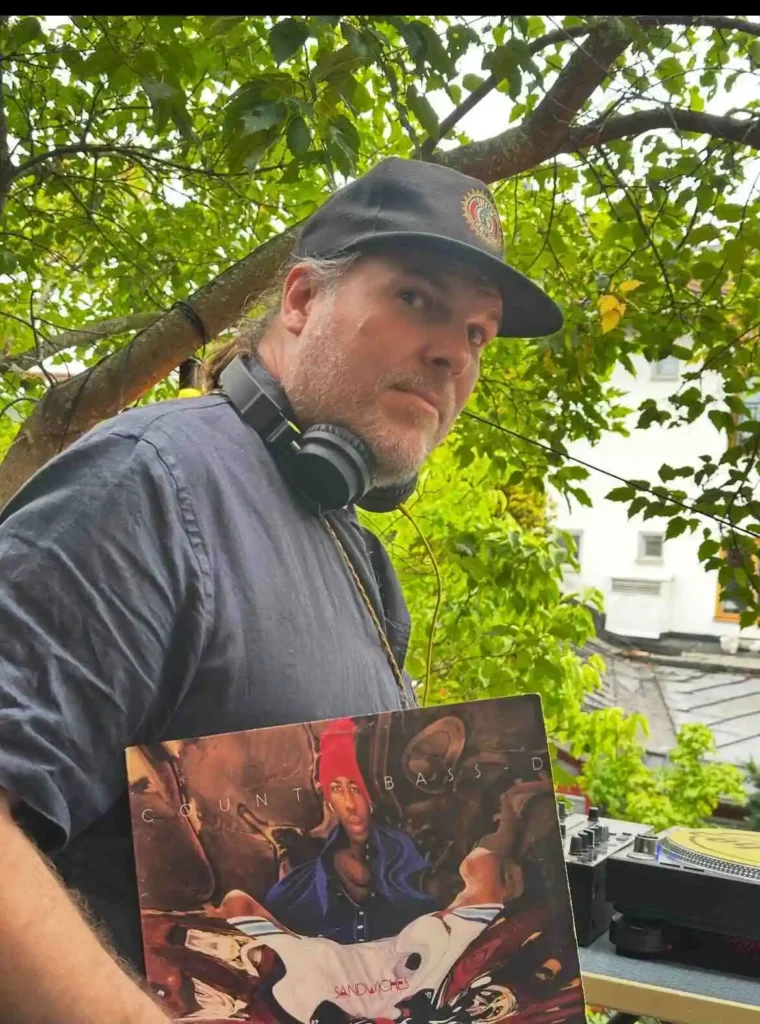DJ Grass står vid ett DJ-bås utomhus med hörlurar runt halsen och visar upp vinylskivan Sandwiches av Count Bass D på Boom Bap Spot-eventet.