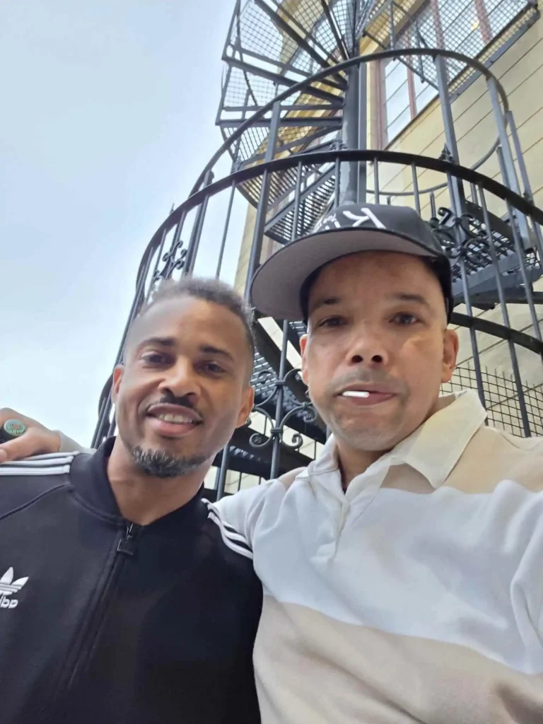 Erik Lundin i svart Adidas-jacka och Ramo från AFC i randig tröja poserar framför spiraltrappan på Mosebacke. – Boom Bap Spot