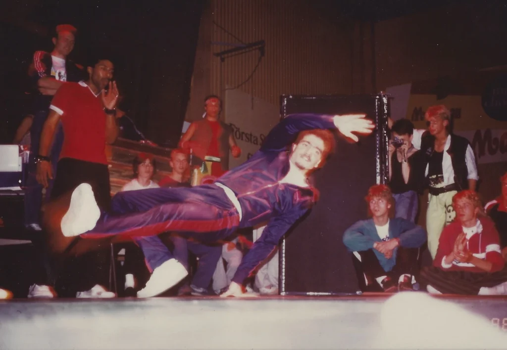 Icey Ice utför ett akrobatiskt breakdance-move på scenen vid Fryshuset i Stockholm, på jamet 2 Def Days of Hipo Hop, omgiven av publik och andra dansare under 1980-talet.