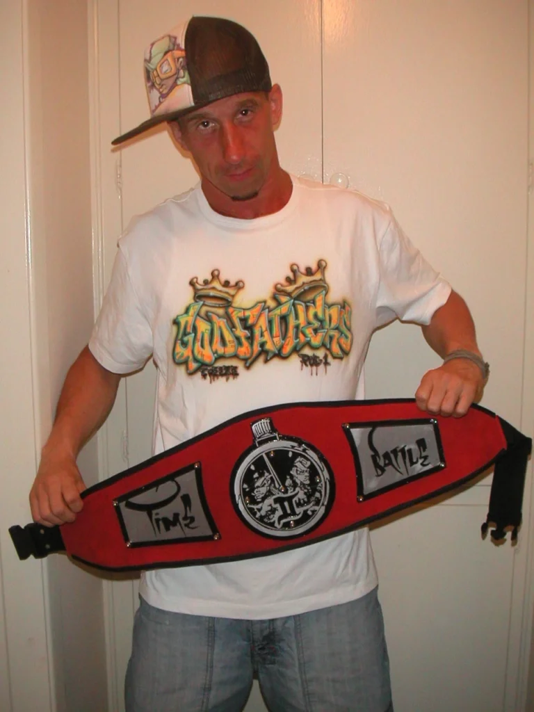 Freeze håller upp mästarbältet från breakdance-tävlingen Time to Battle, iförd en T-shirt med graffititryck och keps.